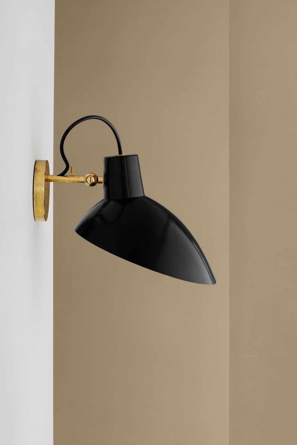 Skråstilt svart vegglampe i metall med messingfeste på hvitt og beige bakgrunn.