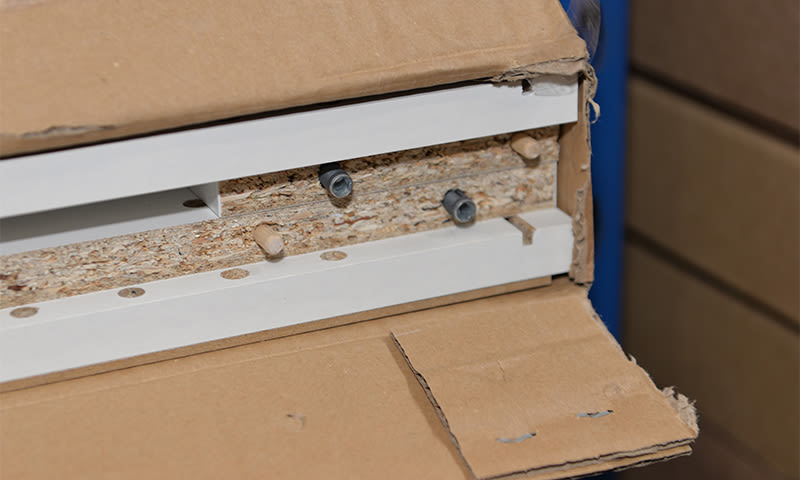 Åpnet brun pappeske med hvite sponplate-møbeldeler, treplugger og metallkamlåser.