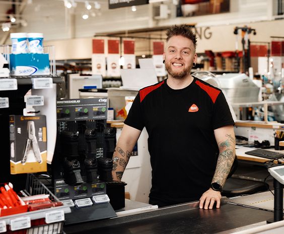 En smilende mannlig ansatt med tatoveringer i svart og rød BM-uniform står ved en kasse i en byggevarebutikk.