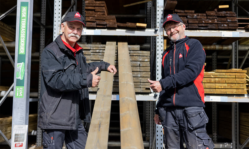 To ansatte i svart-røde jakker demonstrerer lange terrassebord i byggevarelager
