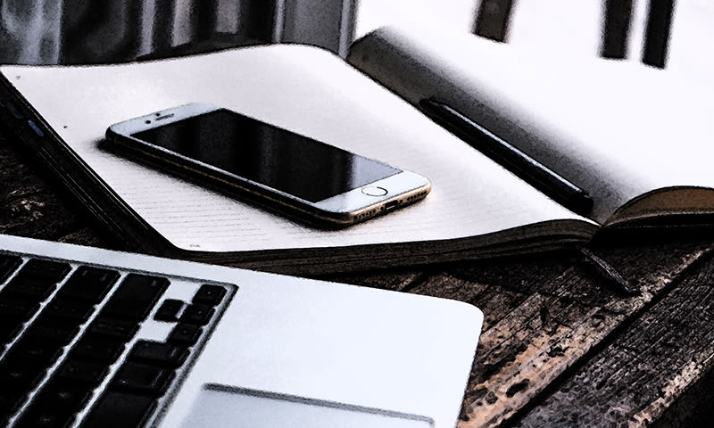 Smarttelefon på åpen notatbok med penn, laptop på rustikt trebord.