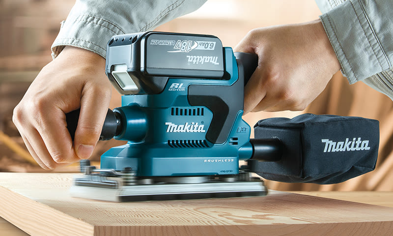 Blå Makita excentersliper i bruk på treplate, to hender holder verktøy og støvpose synlig.