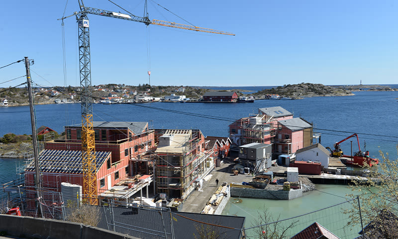 Byggeplass ved sjø med røde hus under oppføring, gult kran, turkis vann og blå himmel
