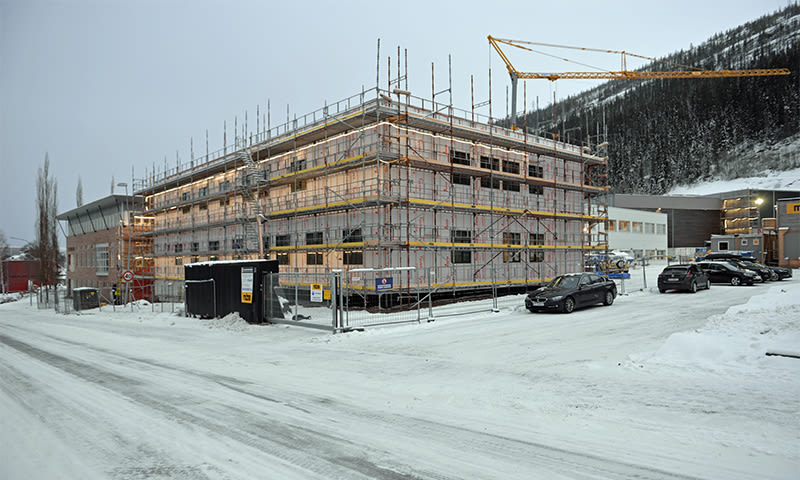 Byggeplass i vinter: nytt trebygg i stillas, gul kran, grå himmel, snø og biler.