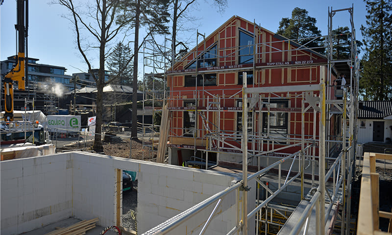 Byggeplass med rødt hus i stenderverk og stillas, hvit grunnmur og gul kran under blå himmel