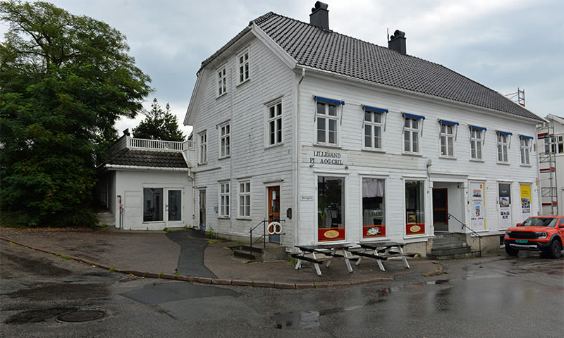 Hvit trebygning med blå markiser og Lillesand Pøl & Grill-skilt, utemøbler og rød elbil