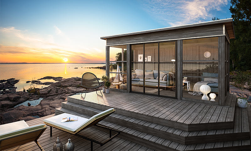 Lite moderne tre- og glashytte på svaberg ved sjø, terrasse med solsenger i oransje solnedgang