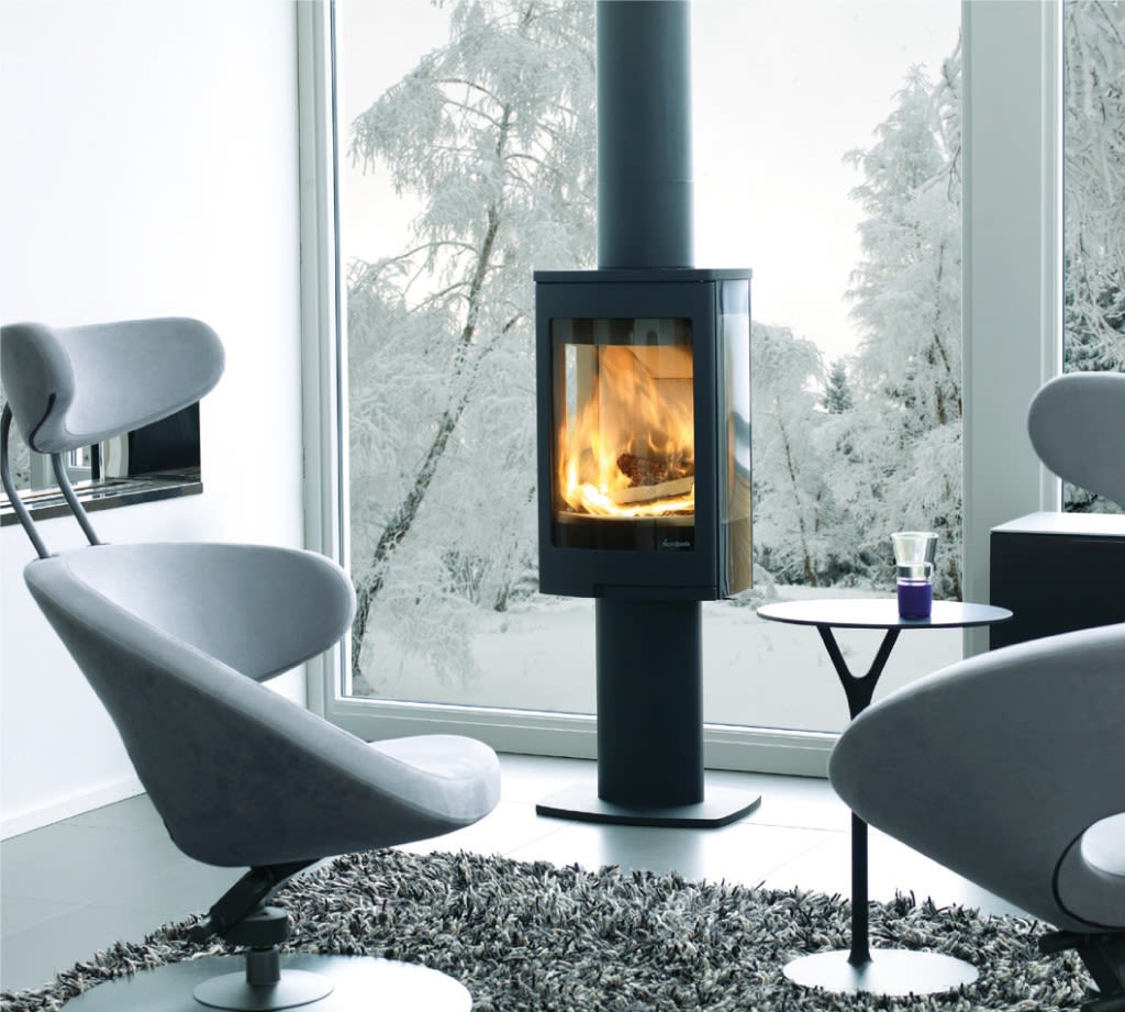 Moderne stue med svart vedovn foran stort vindu mot snødekte trær, grå stoler rundt.