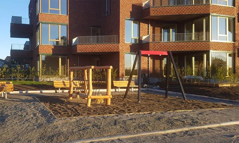 Rødt teglbygg med balkonger, foran mørk jordlekeplass med treklatrestativ og rød huske.