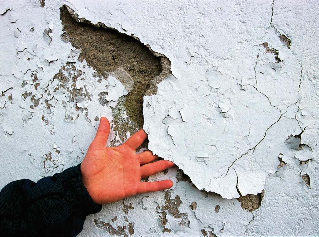 Åpen hånd løfter flassende, sprukket hvitmaling fra rå, brungrå betongvegg.