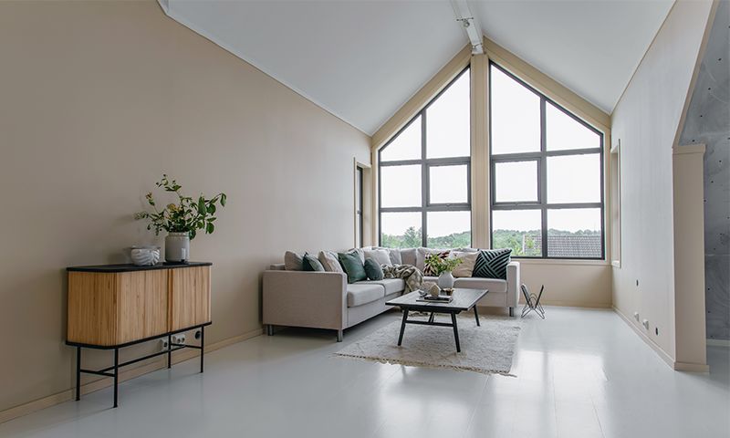 Lys minimalistisk stue med hvitt gulv, beige vegger, hjørnesofa, treskap og store vinduer.