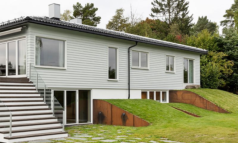 Moderne lysegrå enetasjes hus med svart tak, store trappetrinn med glassrekkverk og gressplen.