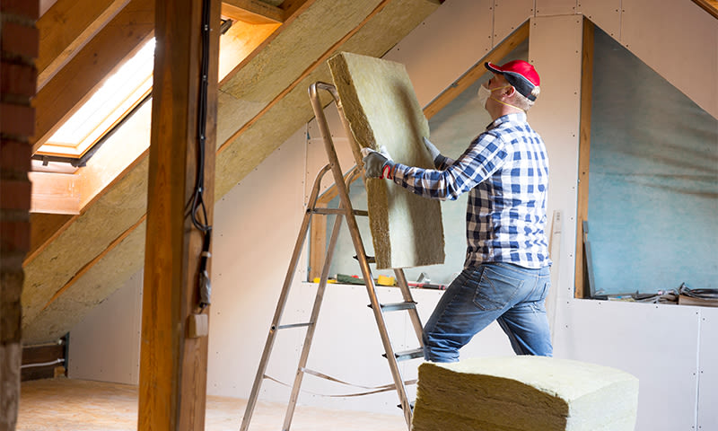 Mann i rutet skjorte, rød caps og munnbind setter opp isolasjonsplate i loft med takvindu