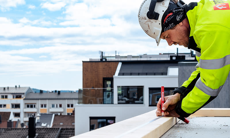 Arbeider i neongul jakke merker trebjelke på tak, byblokker i bakgrunnen
