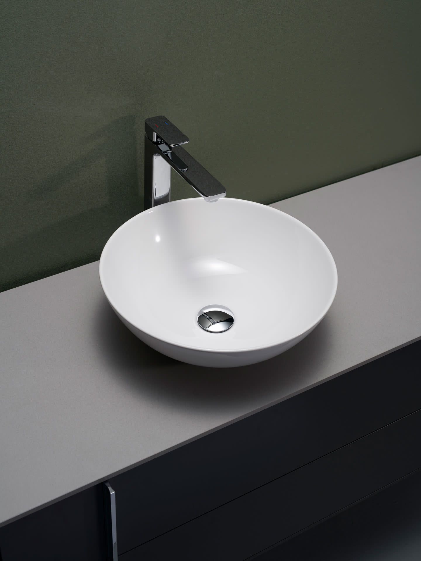 Hvit rund servantskål på grå benkeplate med kromarmatur foran grønn vegg.