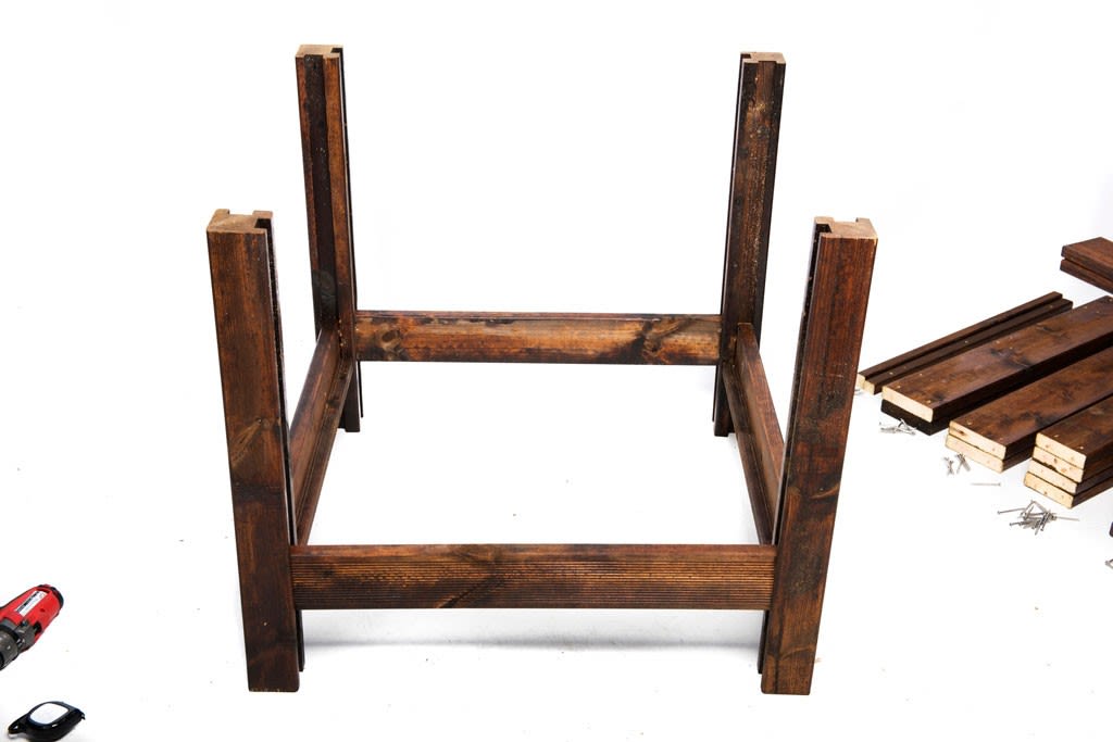 Opp-ned mørkebrunt trebordramme på hvit bakgrunn med løse planker, skruer og rød drill.