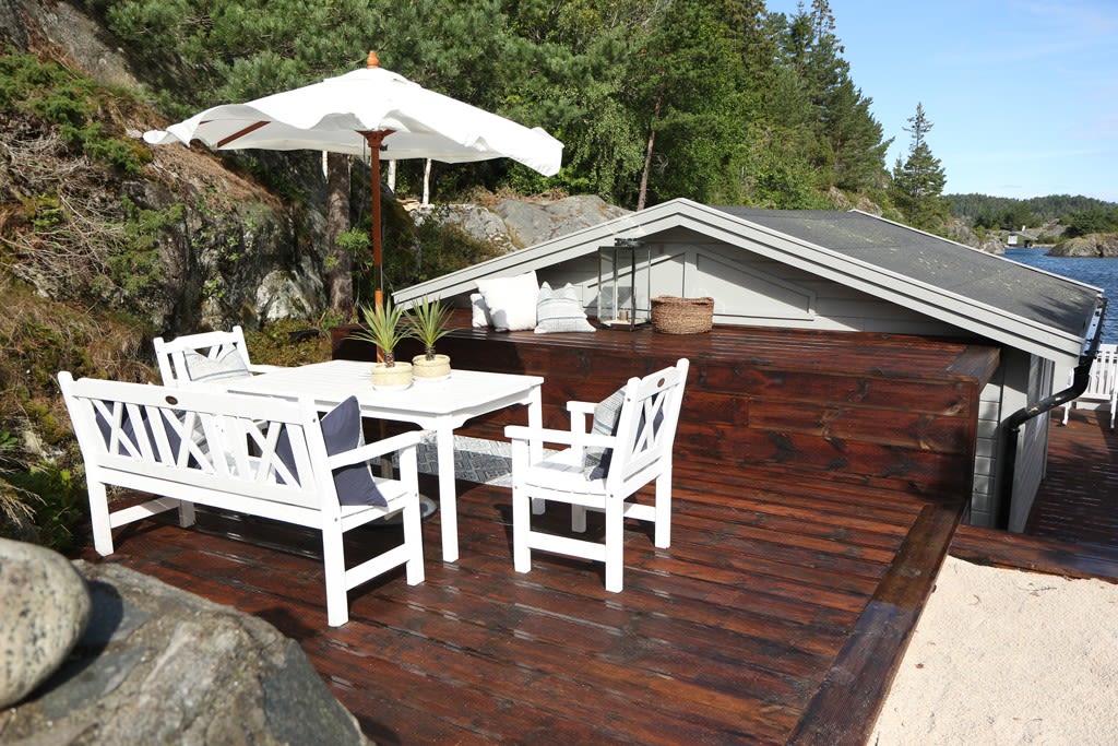 Hvitt utebord med benk og stoler under parasoll på mørk treterrasse ved kysthytte og sjø