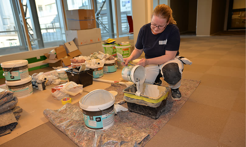 Maler blander hvitmaling i plastbrett på beskyttet gulv med spann og verktøy rundt.