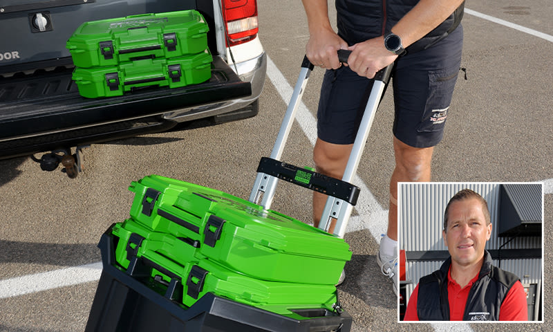 Person drar to stablede grønne verktøykasser på tralle ved hvit pickup på asfaltert parkeringsplass.