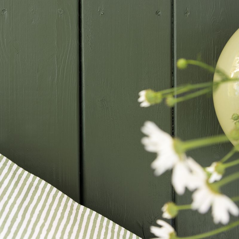 Mørk grønn trevegg med grønnstripet duk, blekgrønn vase og hvite tusenfryd.