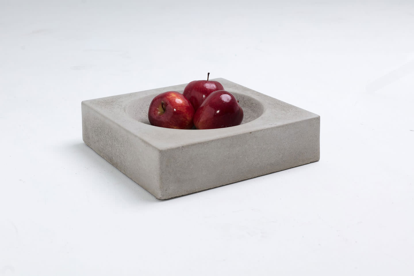 Tre røde epler i en kvadratisk grå betongskål på hvit bakgrunn.