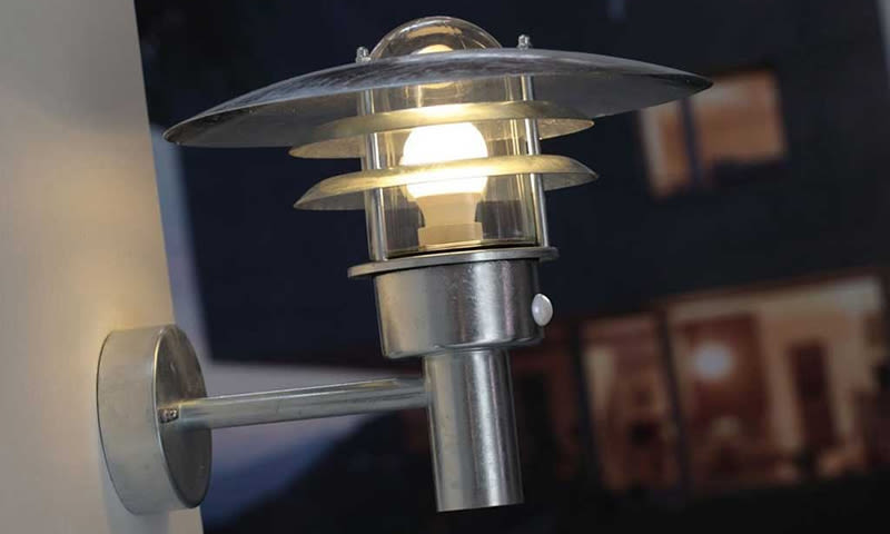 Sølvfarget vegglampe med glasskjerm og lysende pære mot mørk husfasade