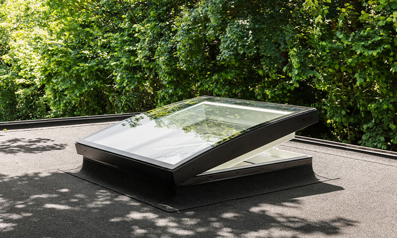 Åpent takvindu med svart ramme på flatt, mørkt tak, reflekterer grønne trær.
