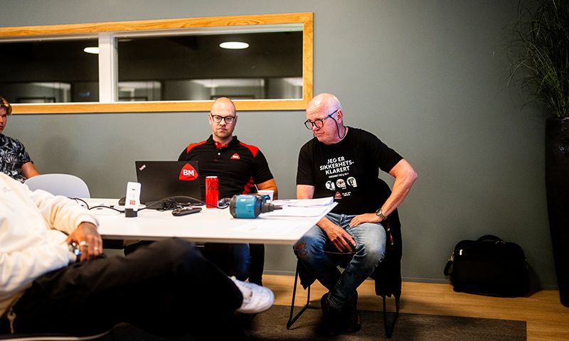 To menn i svart ved hvitt bord med laptop, rød brus, verktøy og papirer foran grå vegg.