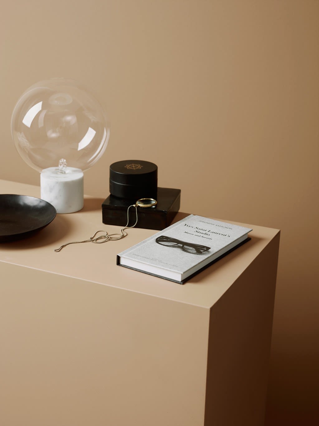 Beige bord med glasskuppel-lampe på marmorfot, svart fat, sorte bokser, gullkjede og bok med briller