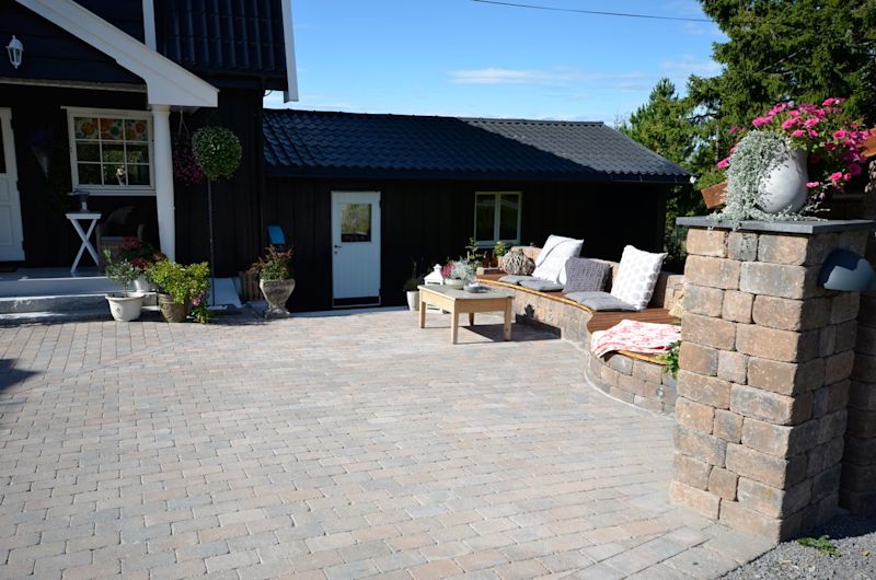 Lyst grå belegningsstein foran svart trehus med buet sittebenk, puter og blomster.