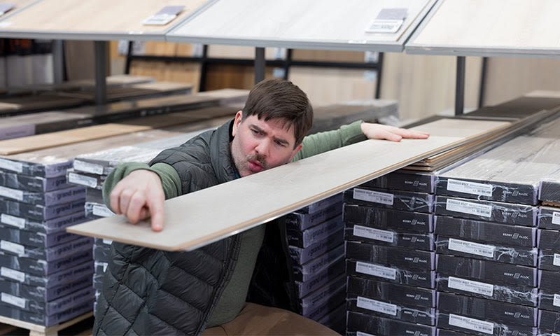 Kunde holder opp lysgrå laminatgulvplanke foran stabler med mørke emballerte gulvplanker.