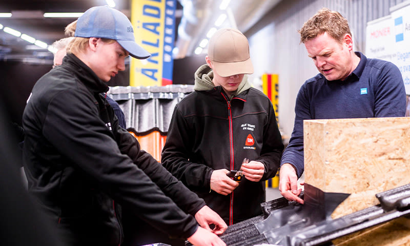 Tre håndverkere i mørke jakker måler sort takpanel i butikkmiljø.