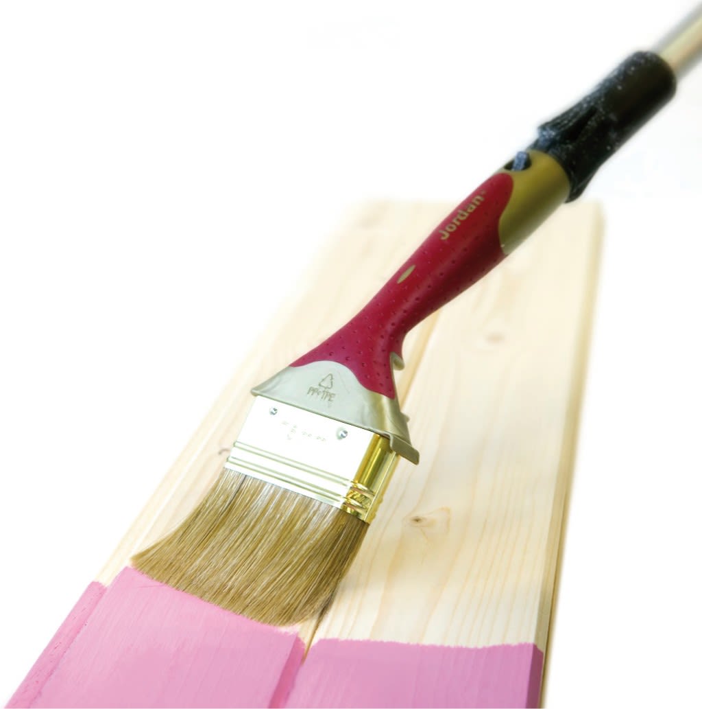 Rød-gul malekost maler en lys treplanke rosa mot hvit bakgrunn.