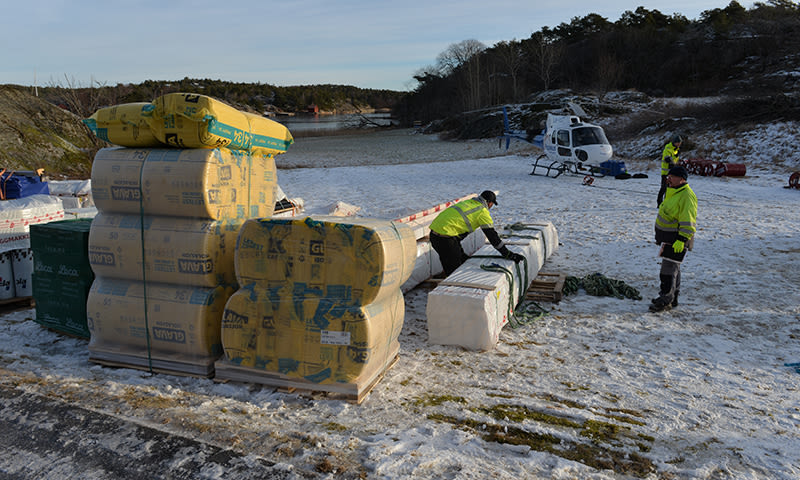 Tre arbeidere i gule jakker laster isolasjon på snødekt strand ved helikopter og skog.