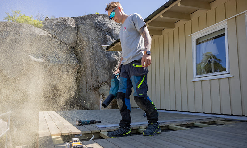 Håndverker blåser støv med varmluftpistol på uferdig terrasse ved lyst hus og granittvegg
