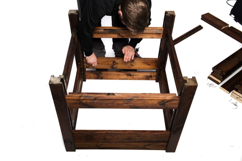 Person i svart bygger mørkebrunt trekasseramme på hvit bakgrunn med planker og skruer
