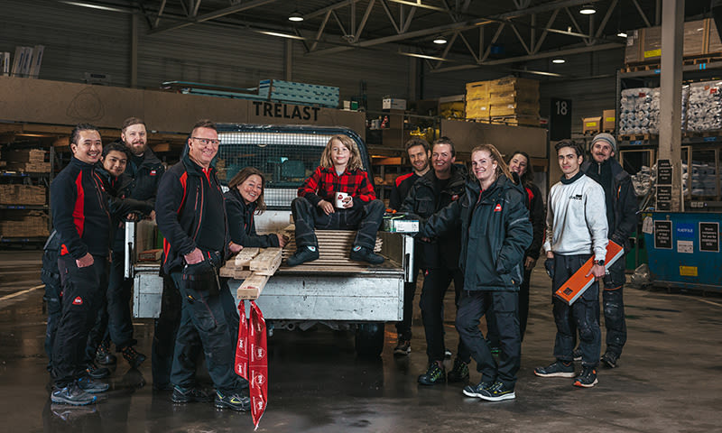 Ti håndverkere i mørke klær står ved en bilhenger med trelast i lagerhall, én i rød rutet skjorte