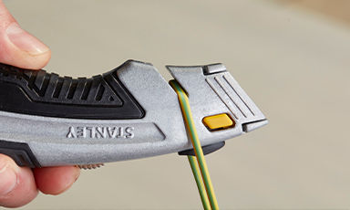 Hånd holder grå Stanley-kniv med gul knapp og kutter grønn-gul elektrisk ledning.