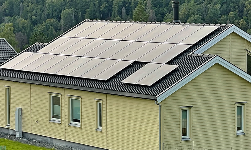 Gult trehus med svarte solcellepaneler på taket foran grønn skog.