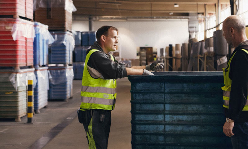 To lagerarbeidere i gule vester løfter stor blå beholder i lagerhall med stabler av plastkasser