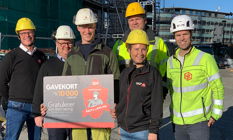 Seks byggearbeidere i verneutstyr holder stort rødt gavekort for årets lærling foran byggeplass.