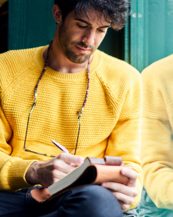 A male author dressed in yellow writing in his story notebook.