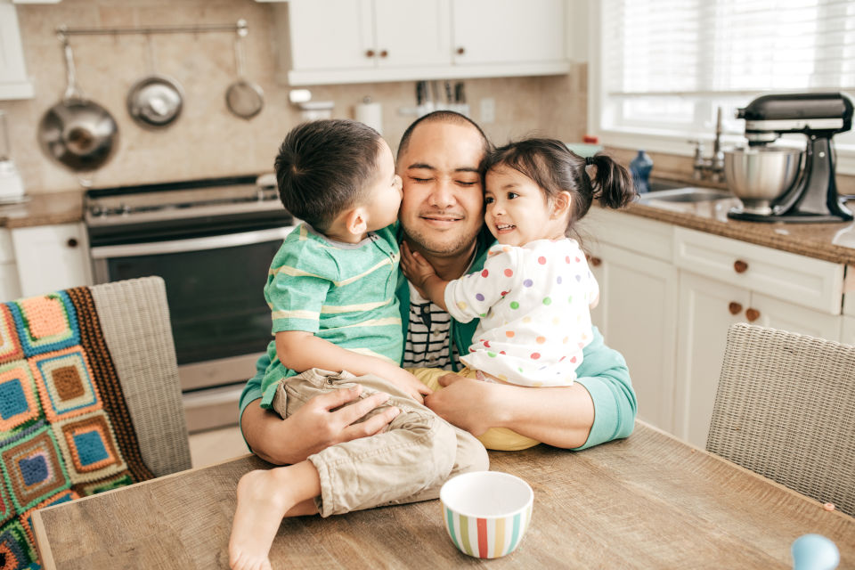 Boy and girl sitting on kitchen table being hugged by father