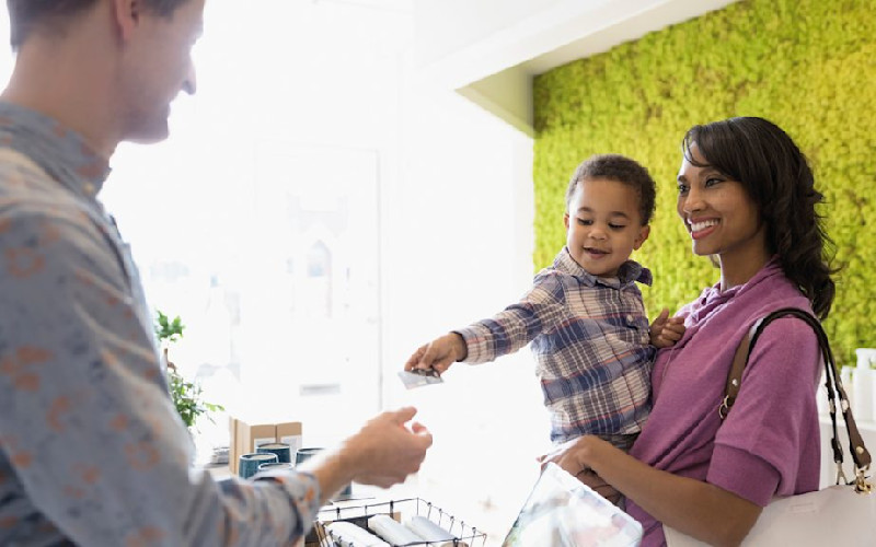 L woman holding toddler handing credit card to cashier