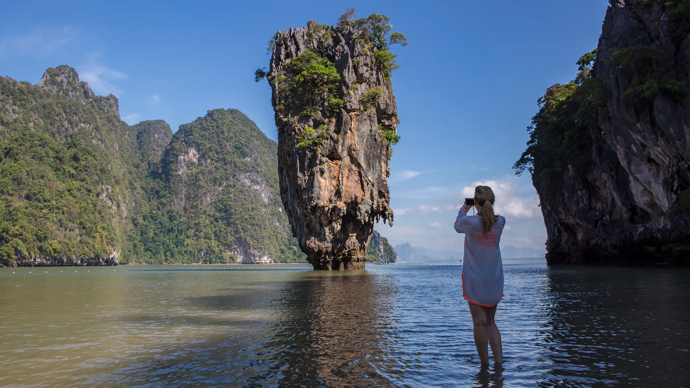 A woman stands in shallow water photographing James Bond Island's iconic limestone rock tower in Phang Nga Bay, Thailand. A woman stands in shallow water photographing James Bond Island's iconic limestone rock tower in Phang Nga Bay, Thailand.