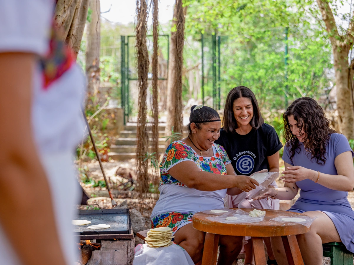 Three women smile while making tortillas outdoors, with a comal over fire nearby, surrounded by lush greenery. Three women smile while making tortillas outdoors, with a comal over fire nearby, surrounded by lush green trees.