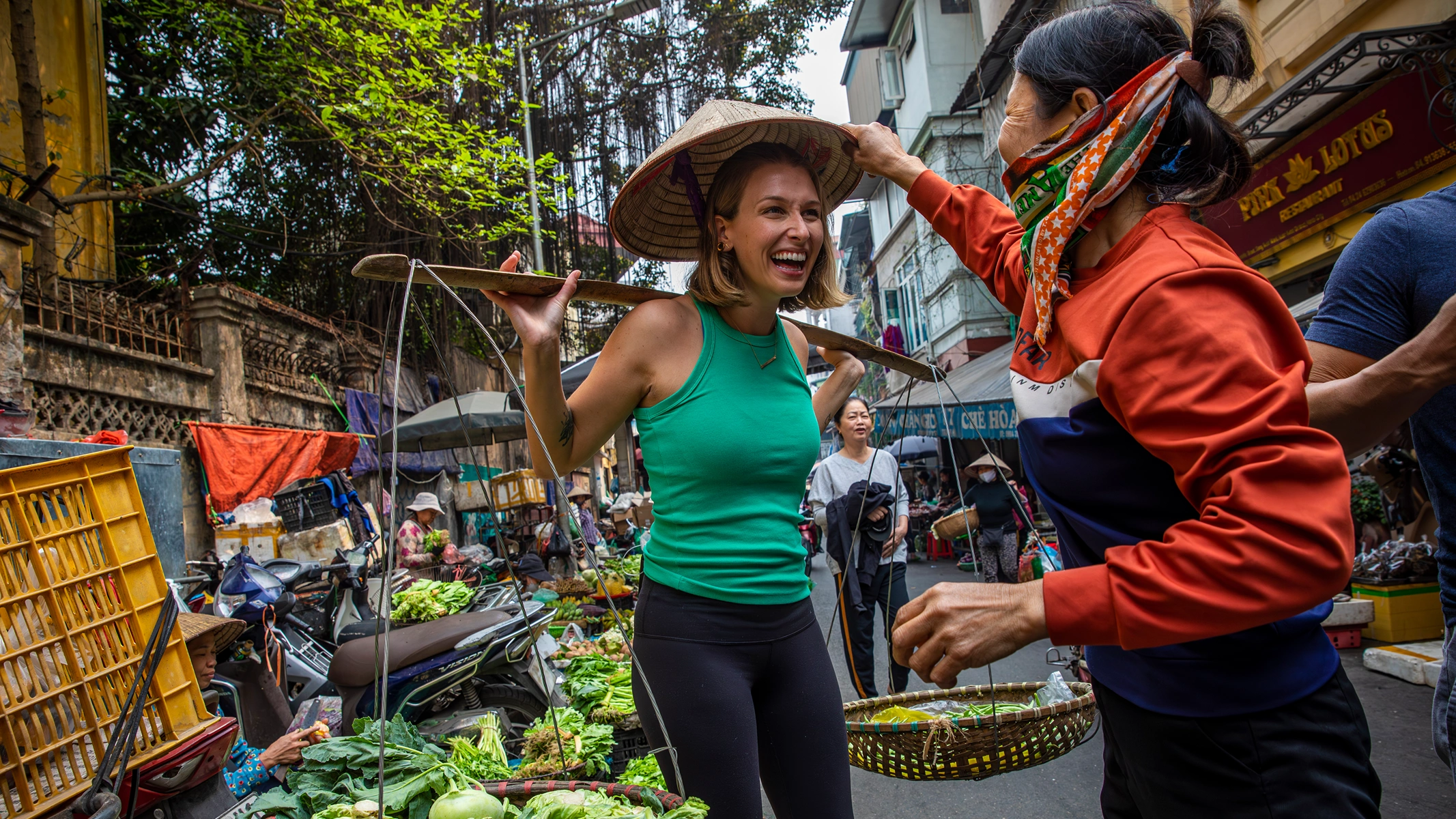 Travel your heart out - Vietnam A smiling woman in a green top tries on a Vietnamese conical hat at a busy outdoor market while a vendor places it on her head.