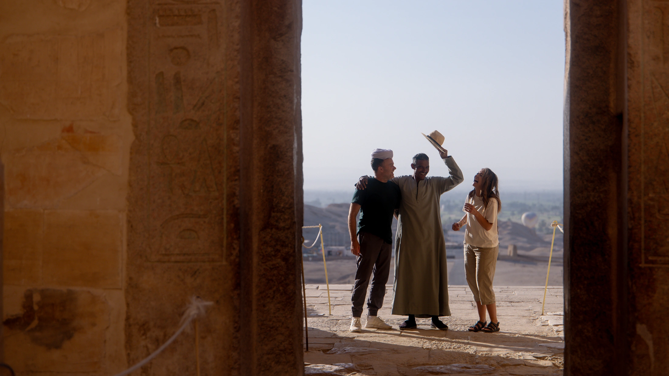 Three people laughing together at an ancient Egyptian temple, framed by hieroglyph-covered stone columns under a clear sky. Three people laughing together at an ancient Egyptian temple, framed by hieroglyph-covered stone columns under a clear sky.