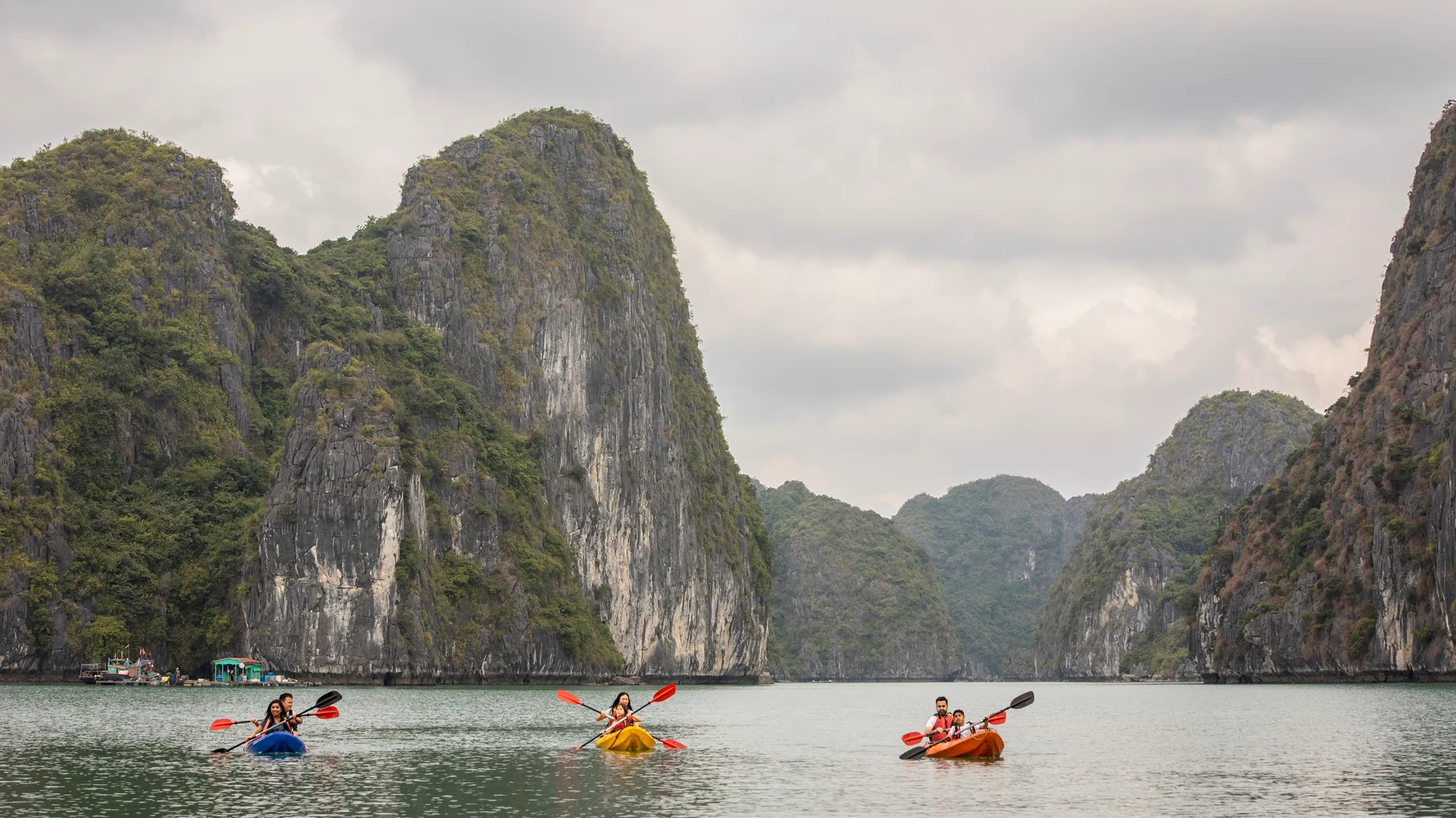 Three kayaks with paddlers navigate calm waters surrounded by towering limestone karsts under a cloudy sky in Ha Long Bay, Vietnam. Three kayaks with paddlers navigate calm waters surrounded by towering limestone karsts under a cloudy sky in Ha Long Bay, Vietnam.