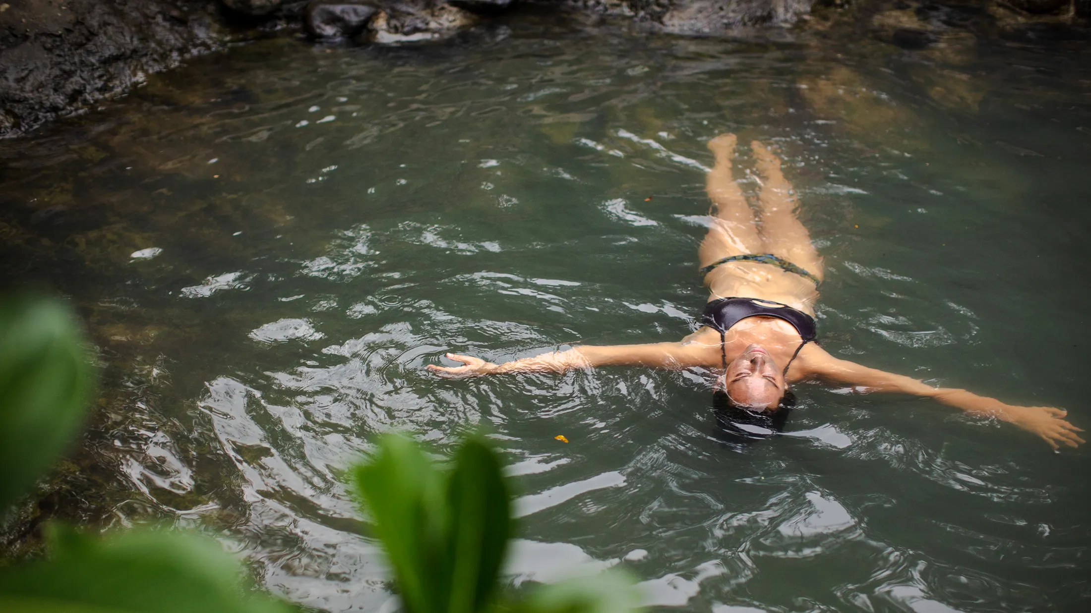 A woman in a black bikini floats peacefully on her back in a calm, green natural pool surrounded by rocks and lush foliage. A woman in a black bikini floats peacefully on her back in a calm, green natural pool surrounded by rocks and foliage.